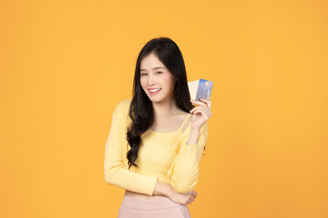 Naklejka premium Cheerful young asian woman in yellow T-shirt holding bank card isolated on yellow background