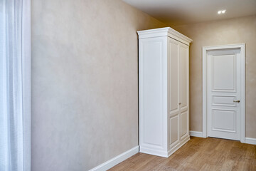White vintage wardrobe with crown molding and white door of classical style in light empty room in luxury apartment