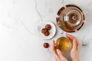 Hand with Glass with green herbal tea  with sweets truffles and teapot on white marble background. Top view. Copy space