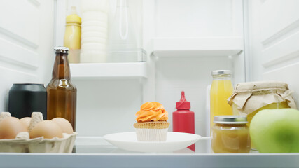 tasty cupcake near eggs, bottle with drinks, jars with honey and fruit puree in fridge.