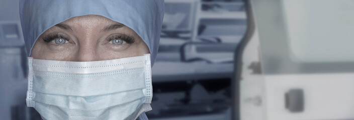 Close-up shot of a female ambulance paramedic in a medical mask looking at the camera against the...