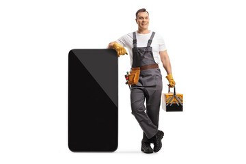 Full length portrait of a repairman in a uniform holding a tool box and leaning on a big smartphone