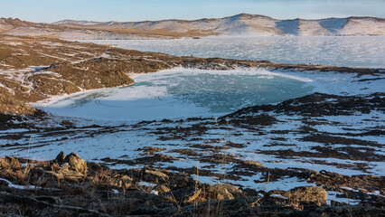 A small frozen heart-shaped lake is surrounded by hills. Round tire tracks are visible on the snow-covered ice. Dry grass on the ground. In the distance - a mountain range against the blue sky. Baikal