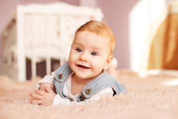 Newborn 5 months old lies on bed on stomach. Baby girl smiles and plays in room. Denim sundress.