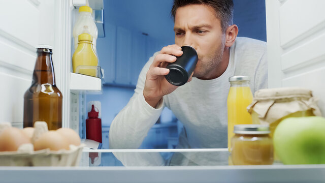 man drinking soda from metal can near fridge with beverages, eggs and bottles with sauces.