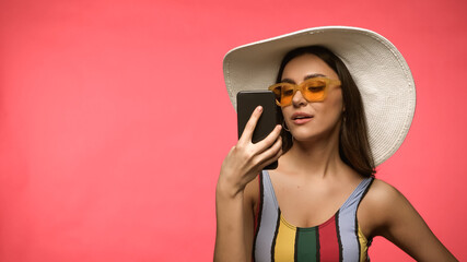 Brunette woman in sun hat and swimsuit holding smartphone isolated on pink.