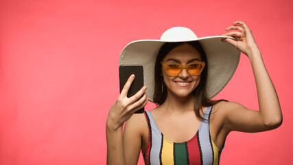 Happy woman in sun hat and swimwear holding cellphone and looking at camera isolated on pink.