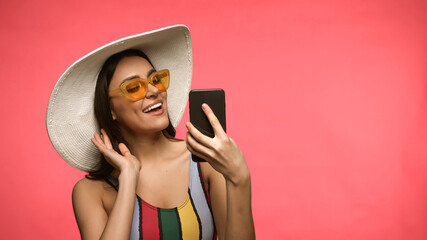 Young woman in sunglasses and swimsuit using smartphone isolated on pink.