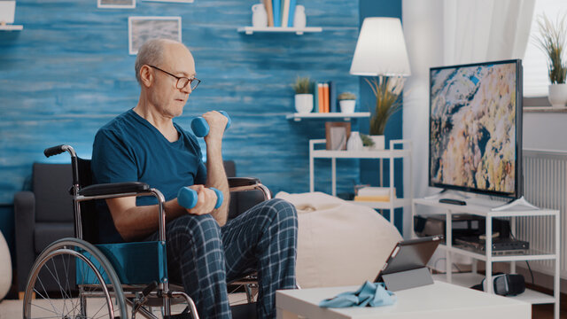 Aged man sitting in wheelchair and lifting weights while following video of workout exercise. Senior person with physical disability using dumbbells and watching training lesson on tablet