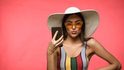 Woman in sun hat and swimsuit holding cellphone and pouting lips isolated on pink.