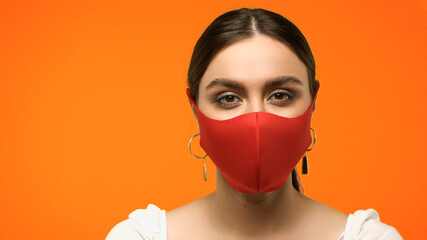 Young woman in protective mask and earrings looking at camera isolated on orange.