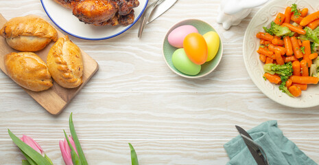 Easter food on white rustic table: pastel colored eggs, roasted chicken and vegetables, buns and spring flowers tulips top view flay lay, Easter family dinner meal with festive dishes, space for text