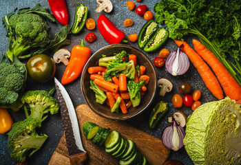 Steamed carrot broccoli salad in bowl and assorted cut fresh vegetables on rustic concrete table,...