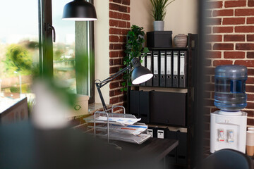 Close up of desk and bookshelf with decorations in office. Empty table with documents and computer, shelves with minimal decor. Water dispenser and bookcase with ornaments and plants.