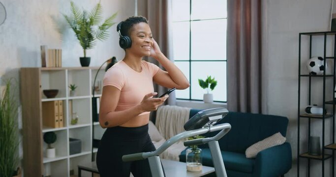 Beautiful smiling sporty slim african american woman in sportswear enjoying favourite music in headphones during home workout on running machine