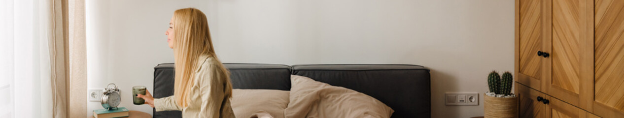 Young blonde woman drinking water while sitting on bed after sleep