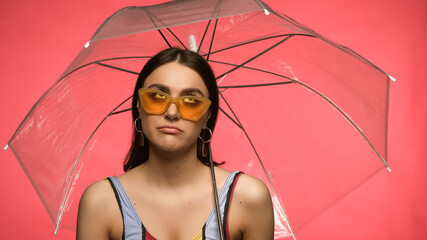 Disappointed woman in swimsuit and sunglasses looking at umbrella isolated on pink.