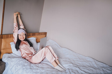 Attractive smiling young woman in pink silk pajamas and eye sleeping mask stretching in bed waking...
