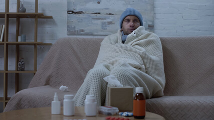 ill man measuring temperature while sitting on couch in warm beanie and blanket near table with...