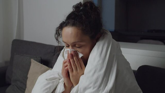 Sick woman coughing, blowing nose, using nasal spray while sitting on couch covered with duvet blanket at home. Adult female with symptoms of flu, cold, virus suffering from seasonal disease indoors