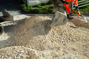 Excavator and roadworker with shovel levelling crushed stone to create flat surface prior placement of asphalt during new road construction