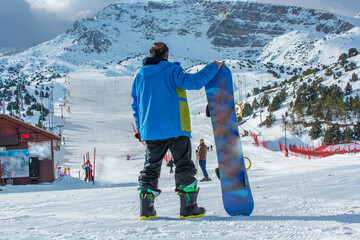 Back view of male snowboarder wearing blue jacket, navy blue pants standing with snowboard in one...