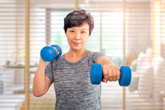 Active Senior Older Asian Attractive Elder Woman Training, Exercising, Workout At Home With Lifting Weights Dumbbells And A Bedroom On A Background. Older Senior Adults Exercise At Home Concept Ideas