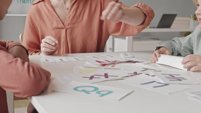 Slowmo shot of two kids learning alphabet at speech therapy lesson with female teacher showing them printed colorful words and letters on cards