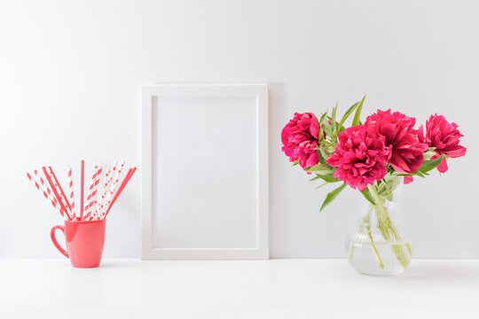 Mockup with a white frame and red peonies in a vase, red paper drinking straws in a cup on a white table