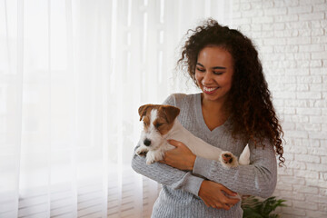 Portrait of young beautiful black woman with her adorable wire haired Jack Russel terrier puppy at home. Loving girl with rough coated pup having fun. Background, close up, copy space.