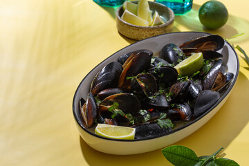 Black mussels in garlic sauce with herbs and lime and two glasses of white wine on the yellow table under the summer sun, copy space for text