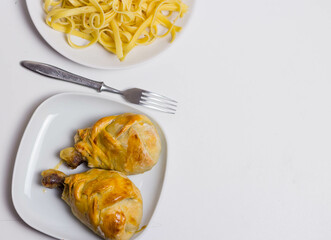 Baked chicken drumsticks on plates and noodles on a plate, fork between plates on white background