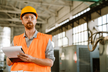 White man wearing helmet and vest writing on clipboard at factory