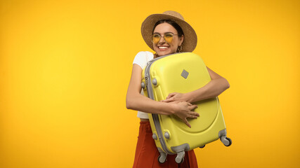 Pretty tourist in straw hat hugging suitcase isolated on yellow.
