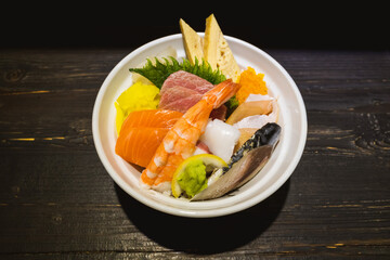 Top view of Sashimi Don, Danburi or Japanese rice bowl with various raw fresh slice of fish, shrimp, squid and tamago egg, on the wooden dark counter.