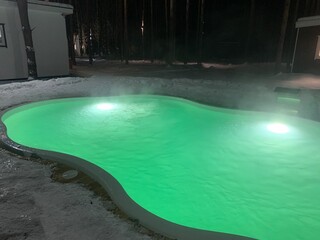 Thermae - a heated outdoor pool illuminated in green at night in the snow with steam coming from...