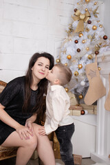 Atmosphere of coming holidays. portrait of happy single mother with her little pleasant boy in white bright room near christmas tree in golden tones.