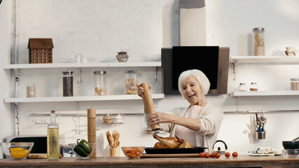 joyful senior woman seasoning roasted turkey near fresh vegetables and oil in kitchen.