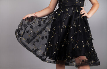 Stylish young woman with black dress, holding a skirt, on gray background.