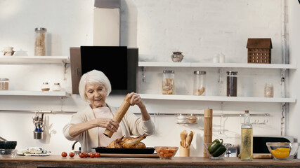 smiling senior woman seasoning roasted turkey near fresh vegetables and oil on table.