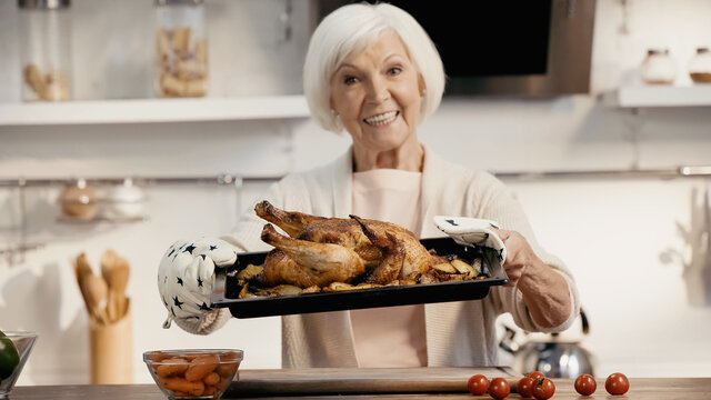 happy senior woman holding oven sheet with delicious turkey and potatoes near fresh vegetables, blurred background.