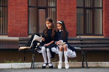 Two schoolgirls is sitting outside together near school building - obrazy, fototapety, plakaty
