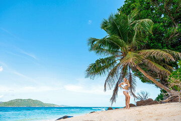 Woman tourist in white swimsuit on tropical beach near palm tree, La Digue, Seychelles