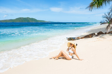 Young beautiful woman wearing white swimsuit and straw hat is sitting on a tropical beach. Summer vacations on picture perfect tropical beach concept.