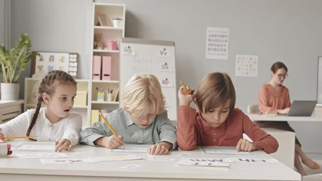 Waist up slowmo shot of three elementary age children sitting at desk in classroom doing test on speech therapy, while their young female teacher working on laptop in background