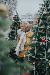 Aged pretty woman with blond hair drinks coffee in the square among the Christmas trees.