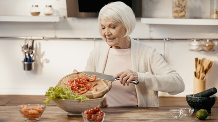 smiling senior woman adding sliced bell pepper to lettuce while preparing vegetable salad.