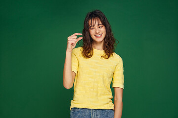 Smiling young girl showing little size gesture with fingers on green studio background. Small...