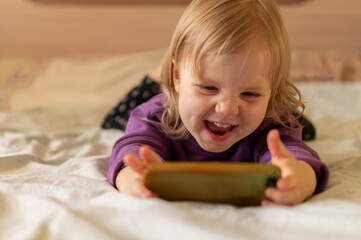 Laughing cute little girl gen Z using online app on mobile phone, browsing internet, playing video games, taking selfie, making call, watching cartoons, resting on bed in bedroom at cozy home.