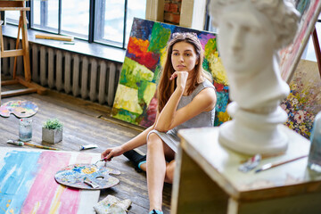 Contemplative female amateur with palette pondering on idea for creative paintings resting in art studio during weekend time, Caucasian woman thoughtful looking away thinking about hobby in workshop